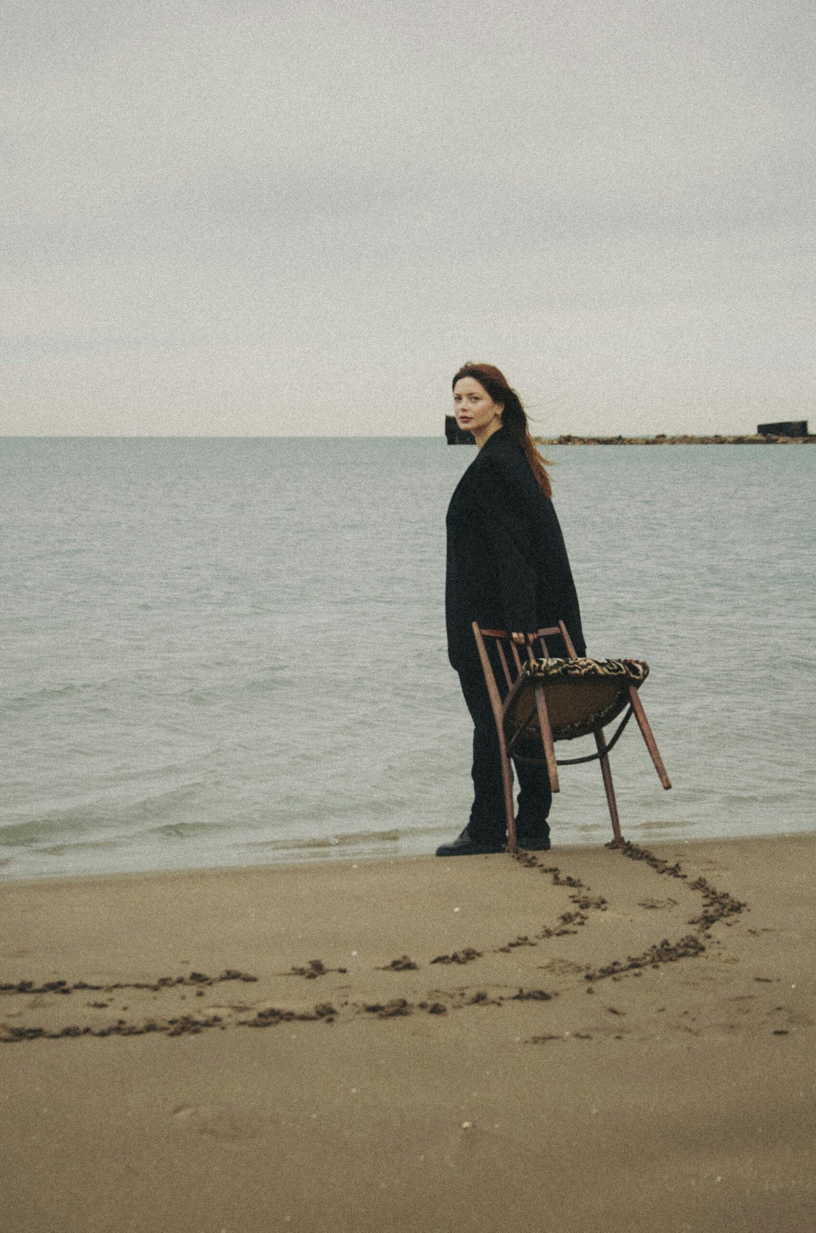 A woman stands alone on the beach in Baku, holding a chair, reflecting solitude.