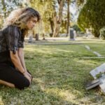 A woman kneeling by a grave in a sunlit cemetery, expressing grief and remembrance.