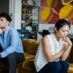 A couple sits indoors on a sofa, both appearing thoughtful and contemplative.